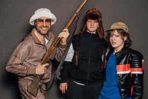 Mann mit Gewehr schaut lächelnd in die Kamera, die zwei Hausptdarstellerinnen stehen verdutzt daneben